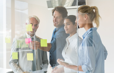 A team is looking at an office wall with sticky notes making up a Kanban process flow